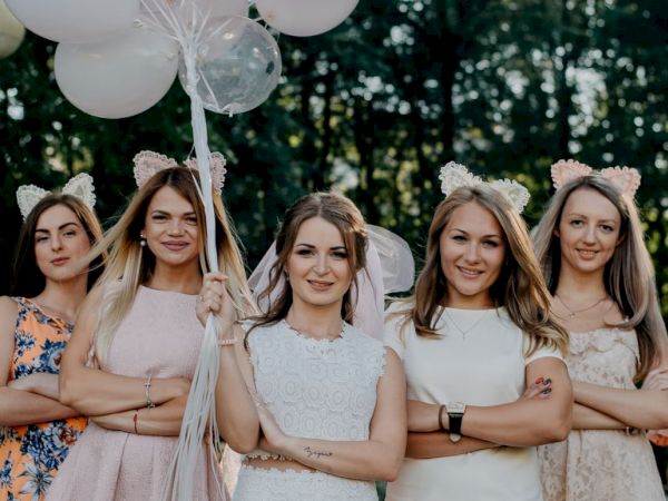 A group of five women, wearing dresses and cat ear headbands, pose outdoors with crossed arms and balloons, smiling confidently.