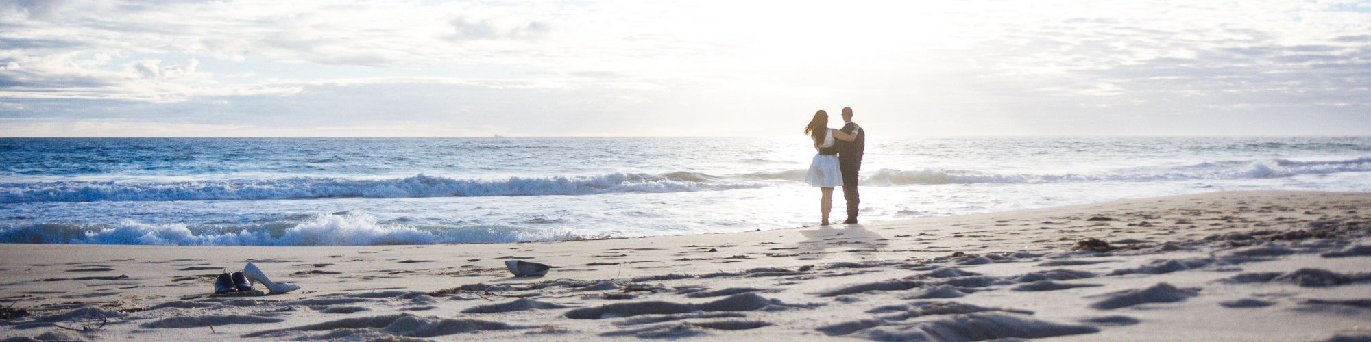 A couple stands on a beach near the ocean, with gentle waves hitting the shore and a clear sky above, creating a tranquil and romantic scene.