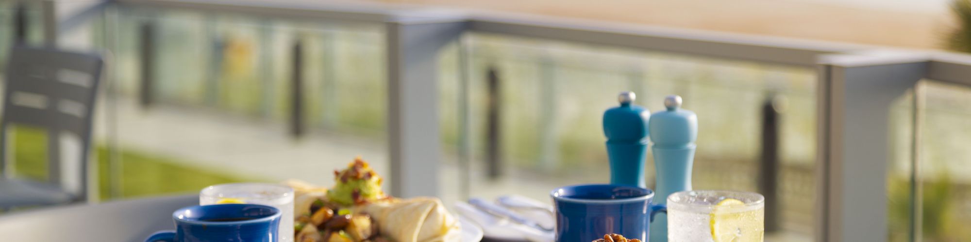 A seaside breakfast setting with pancakes, fruit, blue mugs, cutlery, a glass of water with lemon, and a scenic ocean view in the background.