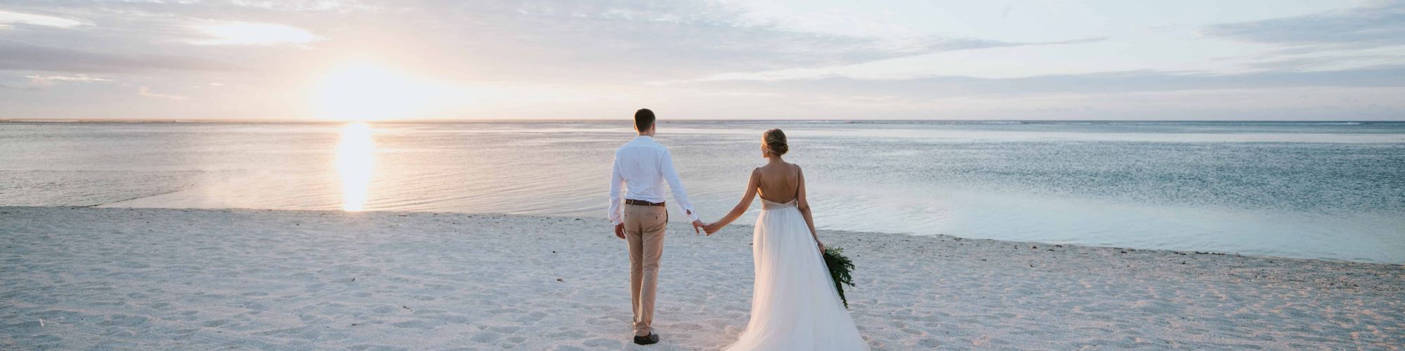 A couple, dressed in wedding attire, stands hand-in-hand on a beach, gazing at a beautiful ocean sunset.