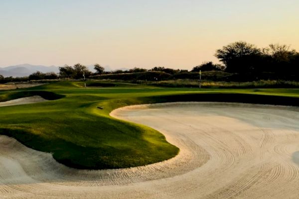 The image shows a golf course featuring a green, well-maintained putting area surrounded by sand bunkers, with trees and a clear sky in the background.