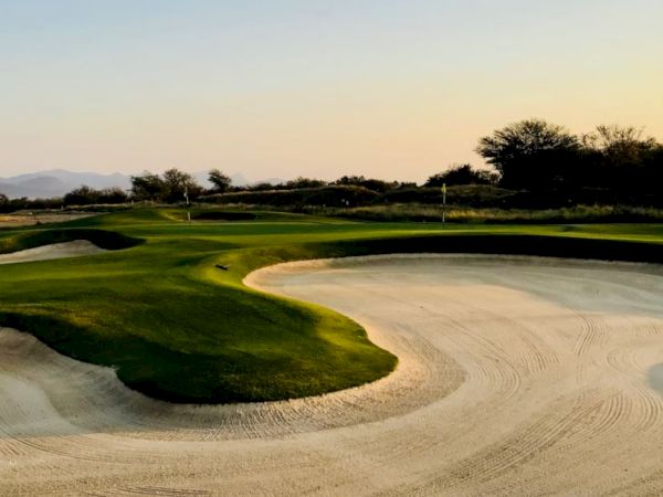 The image shows a golf course featuring a green, well-maintained putting area surrounded by sand bunkers, with trees and a clear sky in the background.