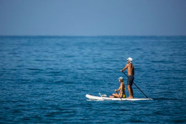 A man and a child are paddleboarding on calm, blue ocean waters under a clear sky. The child is sitting while the man paddles standing up.