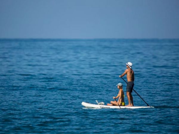 A man and a child are paddleboarding on calm, blue ocean waters under a clear sky. The child is sitting while the man paddles standing up.