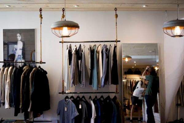 A clothing store with various garments displayed on racks, two people are present, mirrors and hanging lights are seen in the background.