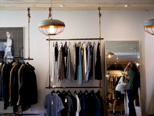 A clothing store with various garments displayed on racks, two people are present, mirrors and hanging lights are seen in the background.