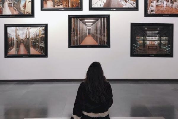 A person is sitting on a bench in an art gallery, observing several framed photographs of different library interiors displayed on the wall.