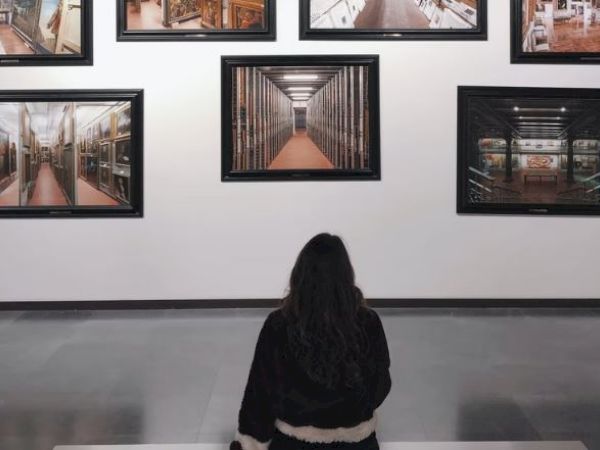 A person is sitting on a bench in an art gallery, observing several framed photographs of different library interiors displayed on the wall.