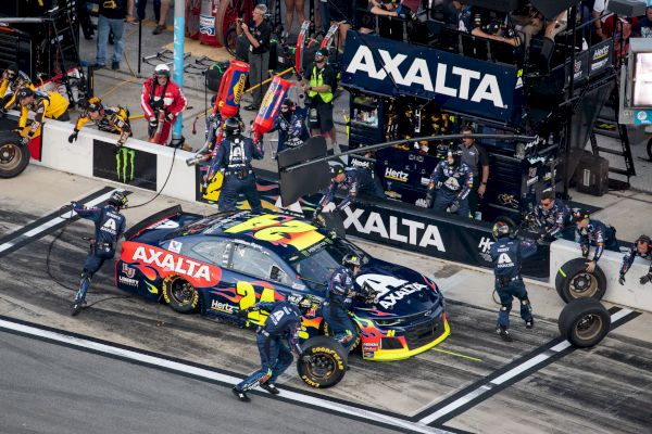 A race car with "Axalta" sponsorship undergoes a pit stop as pit crew members work on the vehicle, with tires and equipment in the pit lane.