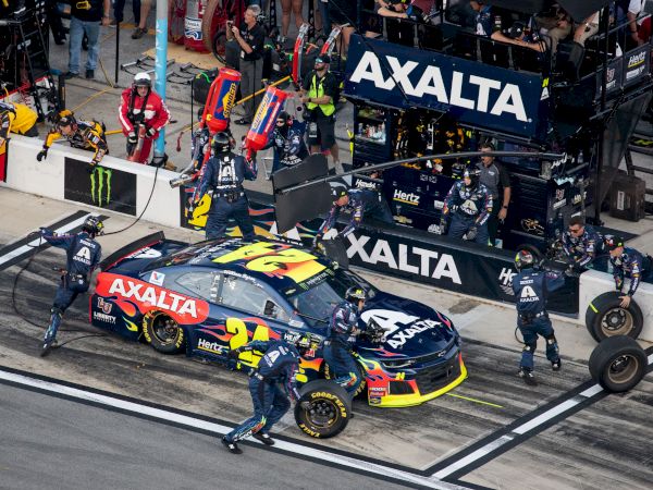 A race car with "Axalta" sponsorship undergoes a pit stop as pit crew members work on the vehicle, with tires and equipment in the pit lane.