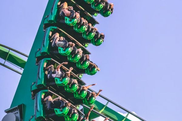 People riding a green roller coaster are in a vertical drop, with their arms raised in the air, against a clear blue sky.