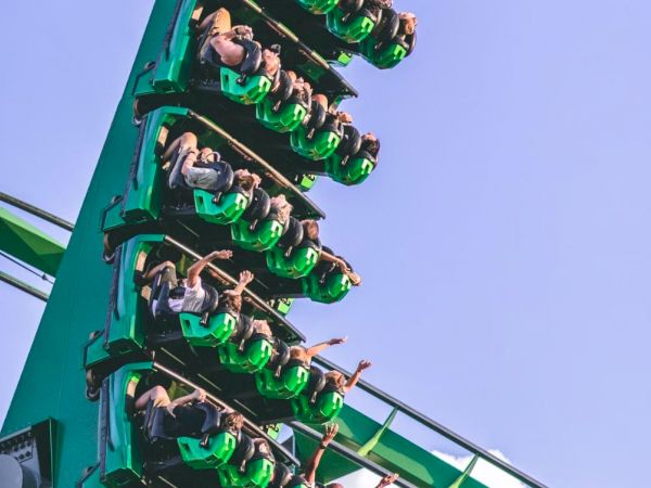 People riding a green roller coaster are in a vertical drop, with their arms raised in the air, against a clear blue sky.