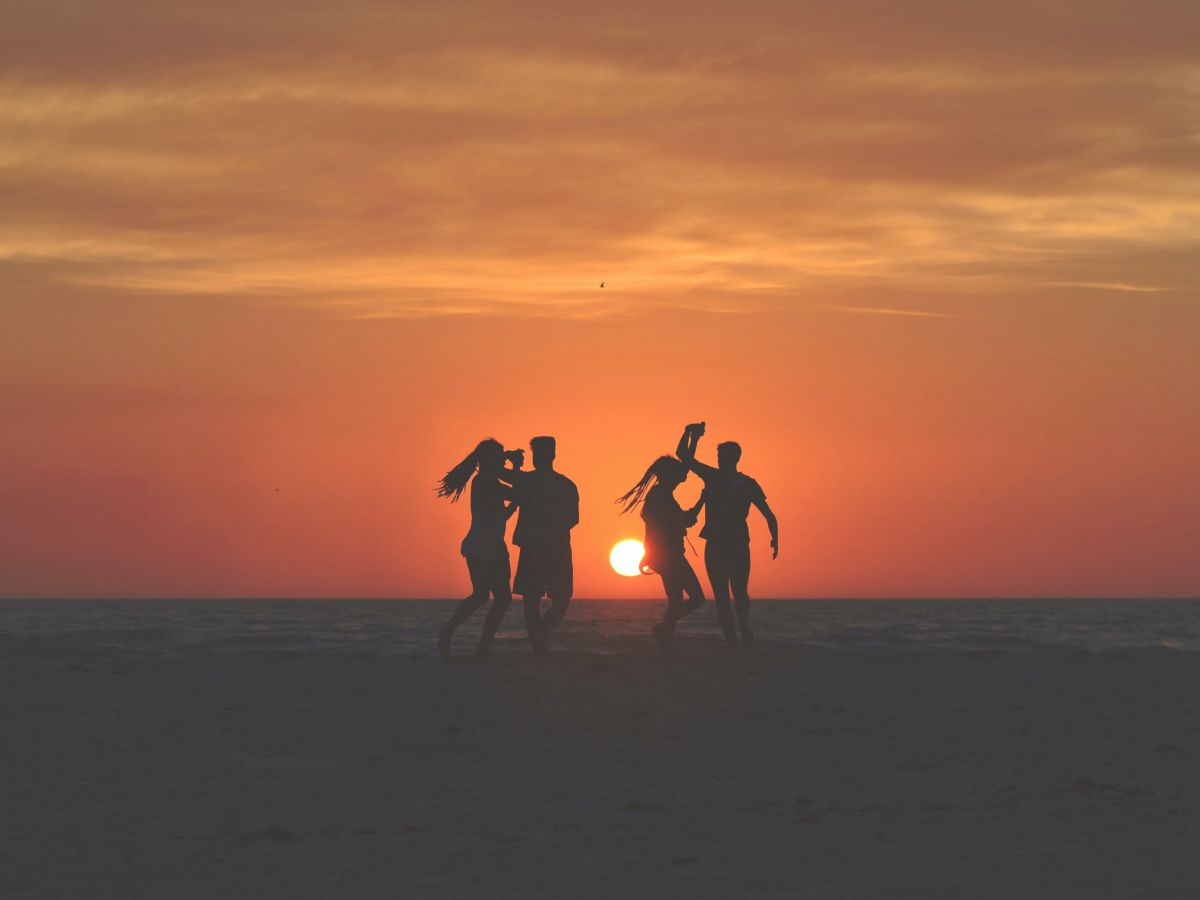 Four people are running on a beach at sunset, with the sun low on the horizon and the sky displaying beautiful orange hues.