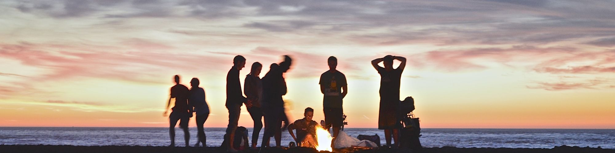 A group of people is gathered around a bonfire on a beach during sunset, with the ocean and a colorful sky in the background.