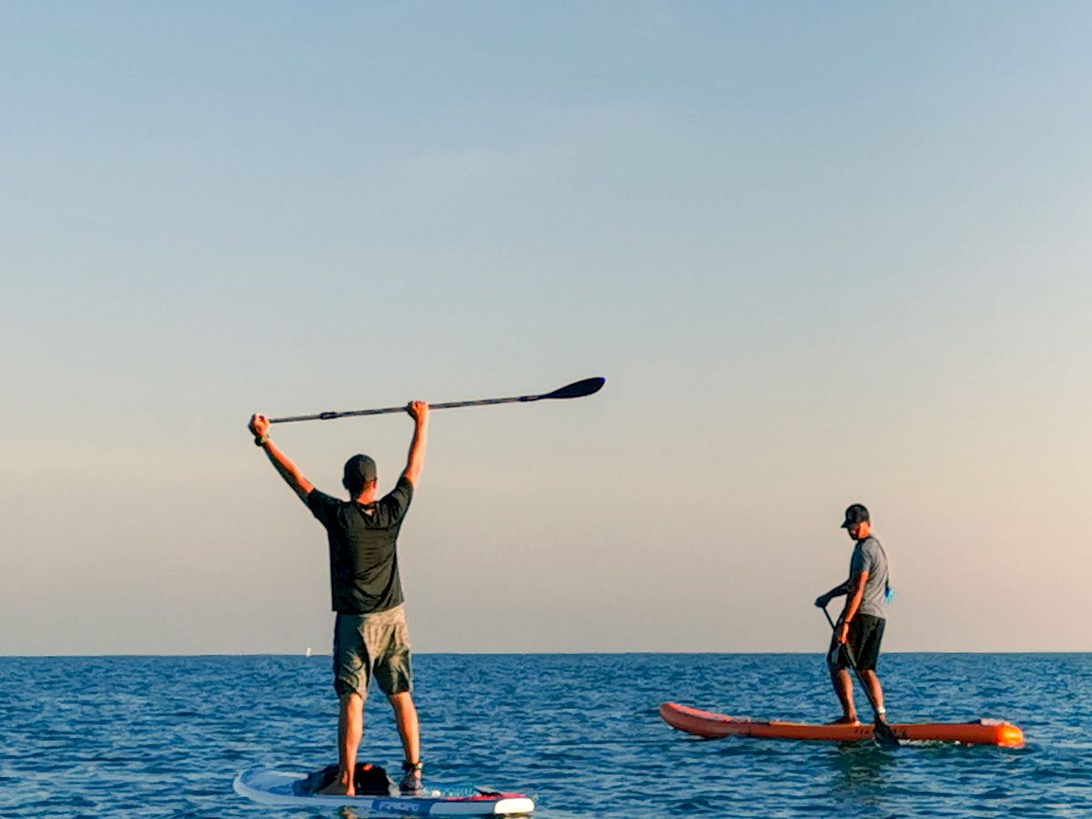 Two people are paddleboarding on a calm sea, holding paddles, with a clear sky in the background. One person has their paddle raised in the air.