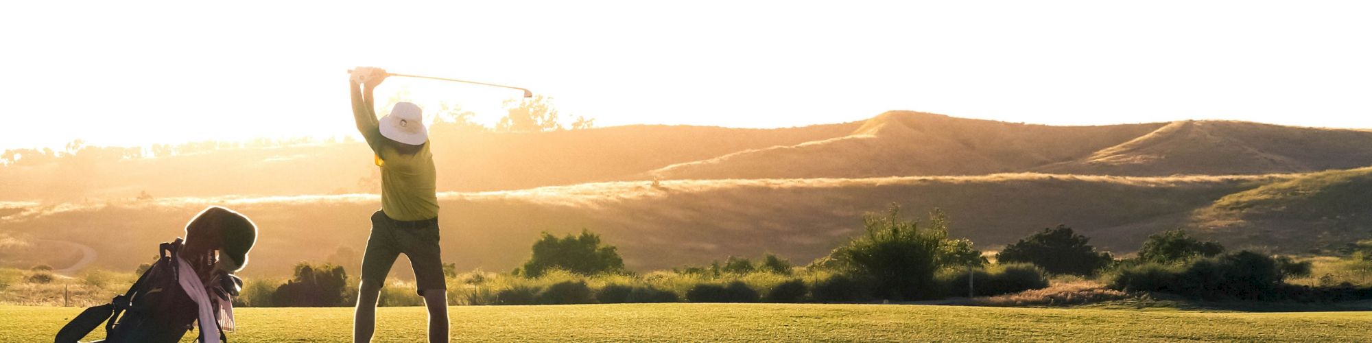 A person is playing golf on a green field during a sunny day, with hills in the background and their golf bag nearby.