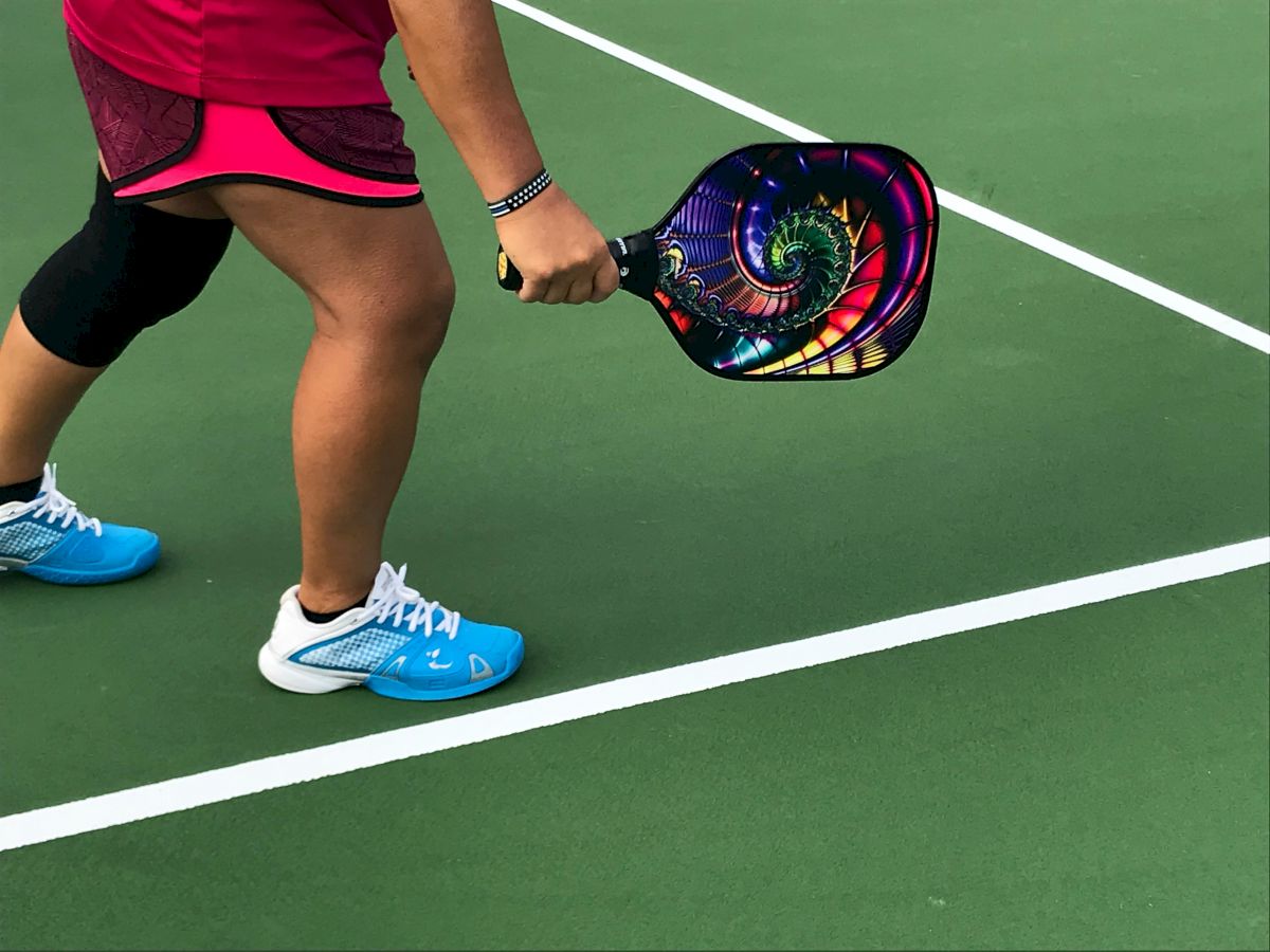 A person is on a court, holding a colorful paddle, preparing to play pickleball. They are wearing blue shoes and sports attire, including a knee brace.