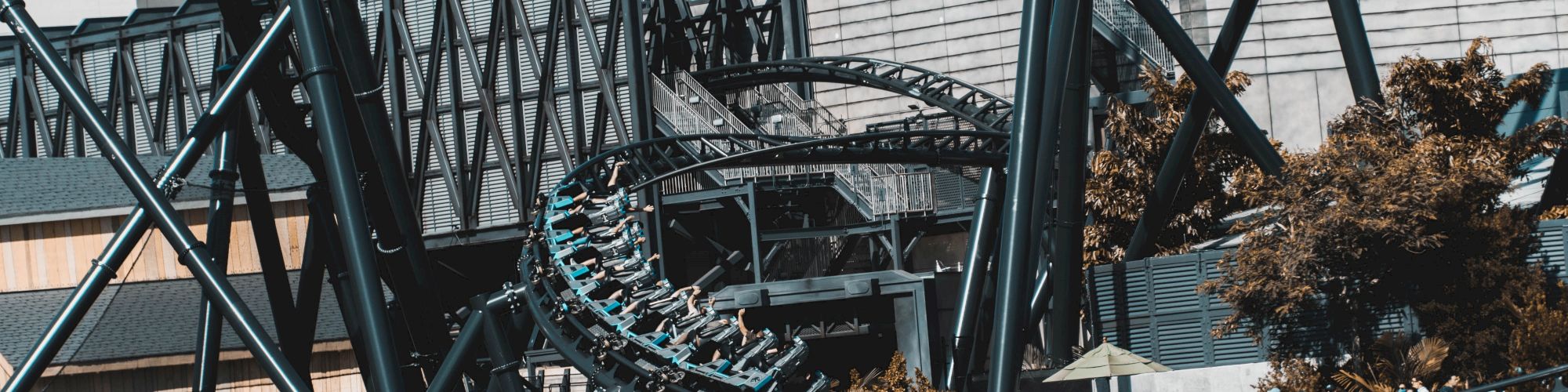 A roller coaster with passengers is seen; it's surrounded by metal structures, buildings, and trees, with water in the foreground.