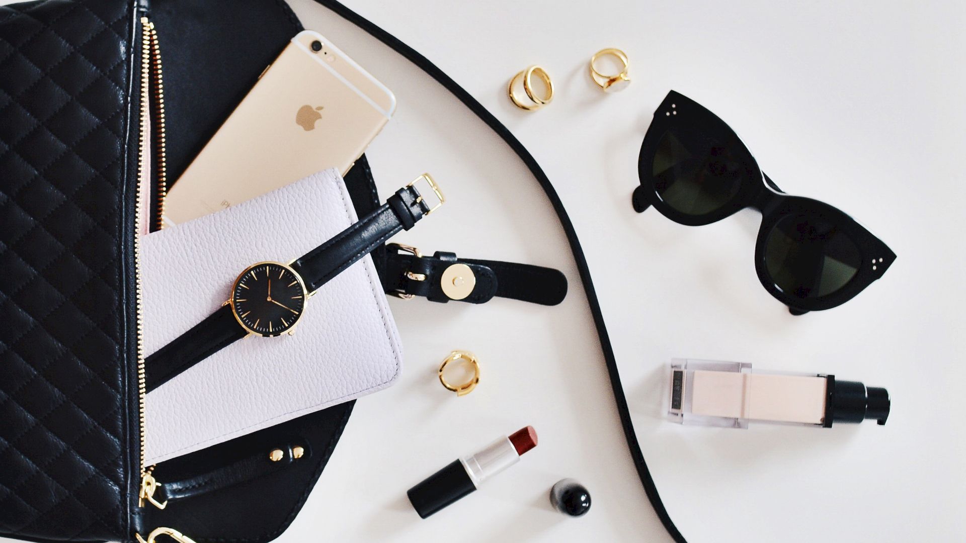A black quilted bag with a smartphone, wristwatch, small notebook, lipstick, rings, sunglasses, and a bottle of nail polish, all laid out on a white surface.
