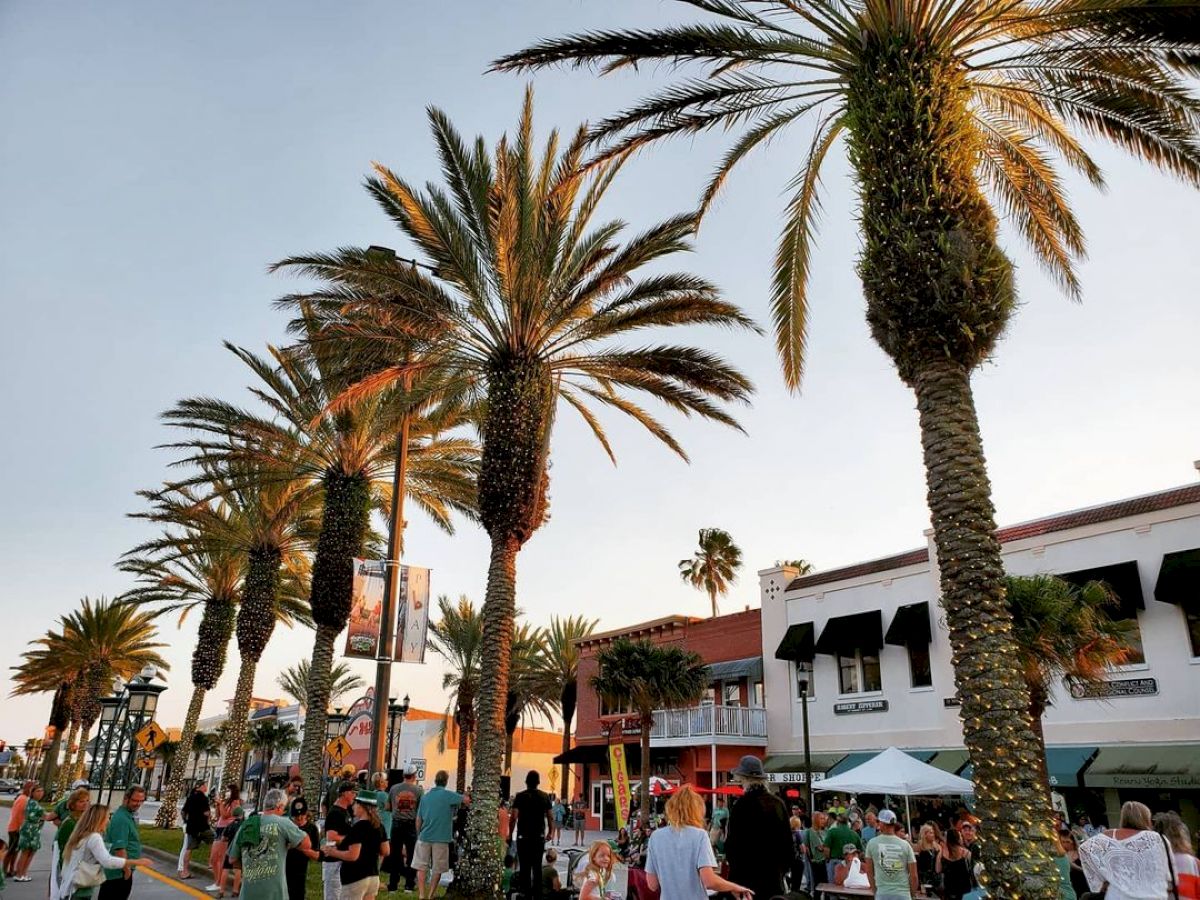 A lively street scene with tall palm trees, people walking, and various shops and buildings in the background under a clear sky.