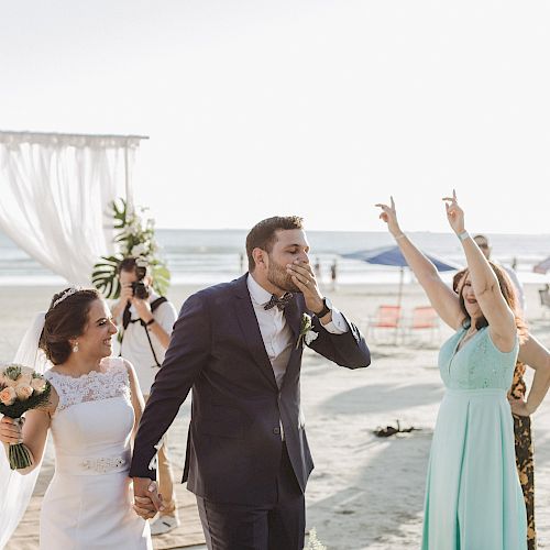 A newly married couple walks hand in hand on the beach, the bride in a white dress and groom in a suit. A bridesmaid cheers in the background.