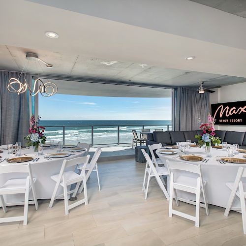 A banquet room with round tables, white chairs, floral centerpieces, and a view of the ocean through large windows, with a sign reading 