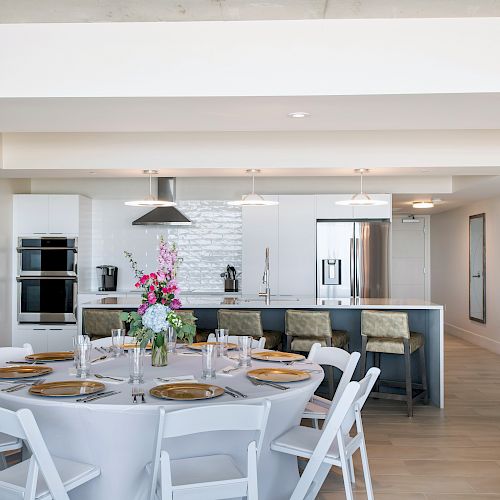 A modern kitchen with stainless steel appliances, a dining table set with a centerpiece and golden plates, and white chairs around it.