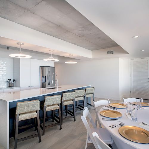 Modern kitchen and dining area with island, bar stools, and table set with golden plates and floral centerpiece.