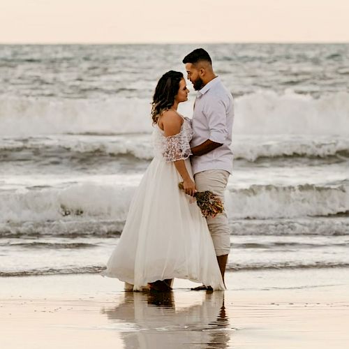 A couple, dressed in elegant attire, stands close together at the beach with waves in the background.