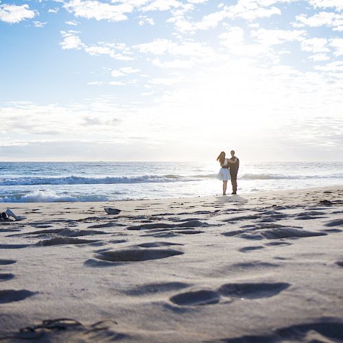 A couple stands on a sandy beach under a bright, cloudy sky, with waves crashing in the background, and shadowed footprints in the foreground.