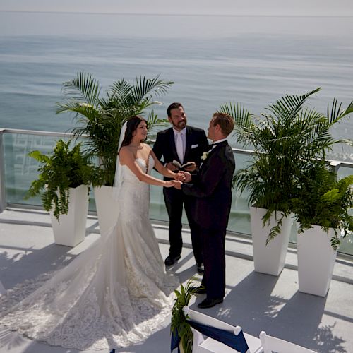 A wedding ceremony is taking place on an outdoor deck with an ocean view, featuring a bride and groom standing with an officiant, surrounded by greenery.