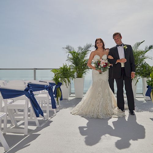 A bride and groom stand at an outdoor wedding venue with ocean views, white chairs adorned with blue ribbons, and green plants in the background.