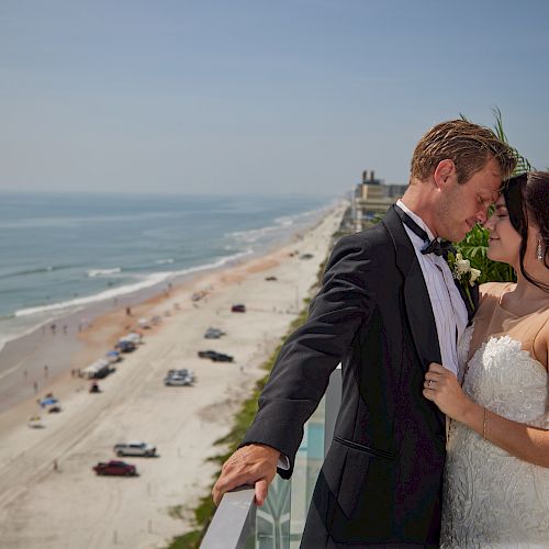 A bride and groom share a moment on a balcony overlooking a beach with cars and people along the shore, against a clear sky.