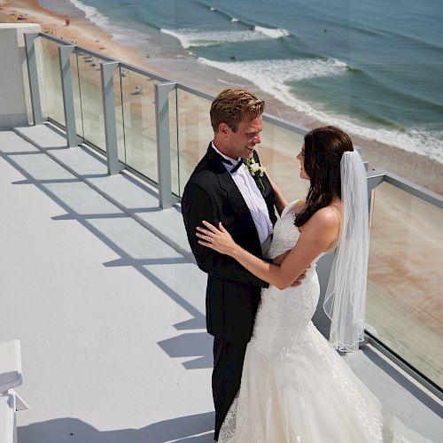 A bride and groom are standing on a balcony overlooking a beach, embracing while wearing their wedding attire. It is a sunny and beautiful day.