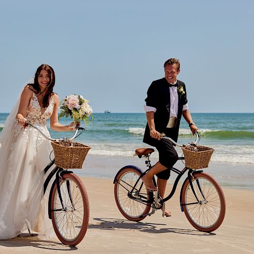 A smiling bride in a white gown and groom in a tuxedo ride bicycles on a sandy beach near the ocean, both with baskets on the front.