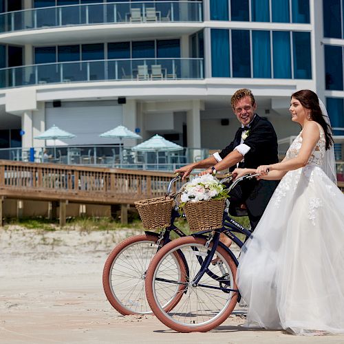 A bride and groom ride bicycles with flower baskets on a beach in front of a modern building, both smiling in wedding attire.