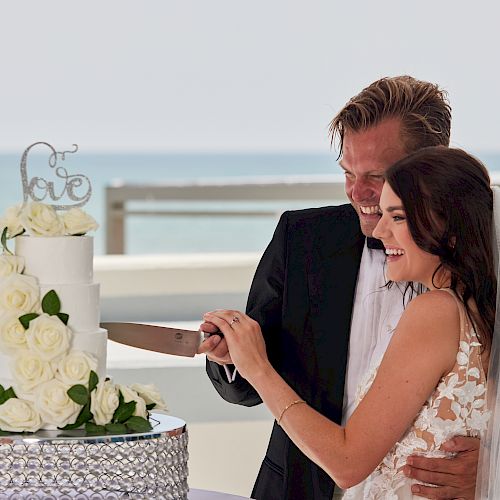 A bride and groom are cutting a white wedding cake adorned with roses, with an ocean view in the background, both smiling happily.