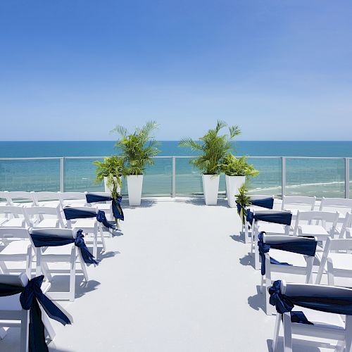 The image showcases a seaside wedding setup with white chairs adorned with navy bows, facing an altar with potted plants under a clear blue sky.