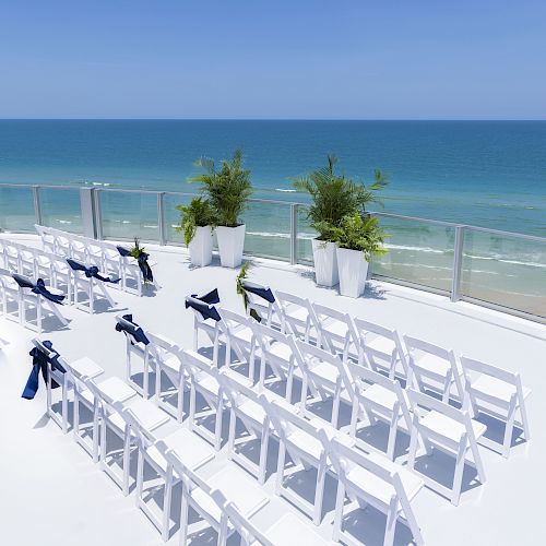 A seaside venue setup with white chairs adorned with blue ribbons, arranged in rows on a terrace with a glass railing overlooking the ocean.