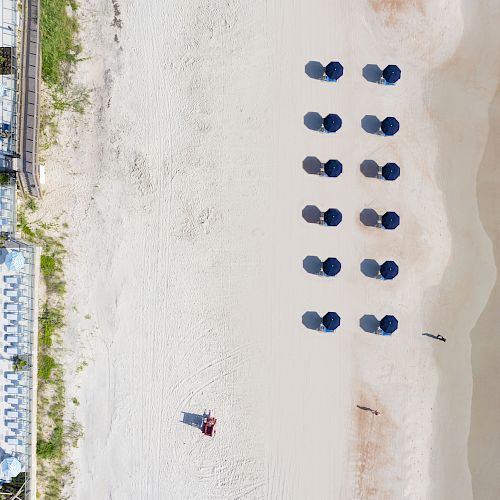 Aerial view of a beach with a row of blue umbrellas, a swimming pool on the adjacent property, and waves reaching the shore.