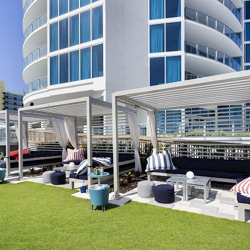 An outdoor lounge area with cabanas, chairs, and grass, beside a modern building with curved balconies.