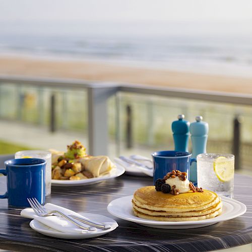 A breakfast table by the beach featuring pancakes, two blue mugs, a glass of water with lemon, and a plate of food, with an ocean view in the background.