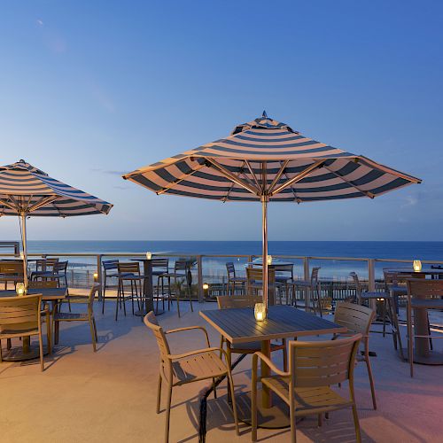 An outdoor dining area with striped umbrellas, tables, chairs, and lit candles overlooking the ocean under a clear blue sky at sunset.