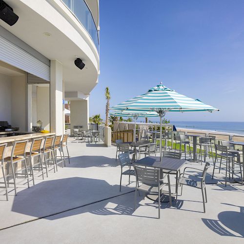 An outdoor beachfront bar and dining area with tables, chairs, a striped umbrella, and a scenic view of the ocean under a clear, blue sky.