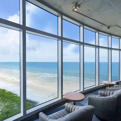 A room with large windows offers a scenic view of a beach and the ocean, featuring comfortable seating and small tables arranged by the windows.