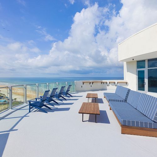 A rooftop terrace with ocean views, featuring blue lounge chairs, a large cushioned bench, and modern glass railings under a partly cloudy sky.