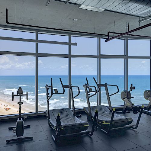 A gym with exercise machines is positioned by large windows, offering a scenic view of a beach and the ocean outside, under a bright sky.