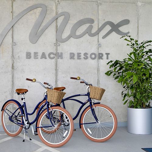 Two blue bicycles with wicker baskets are parked near a potted plant, under the sign "Max Beach Resort", on a patio with a concrete wall.