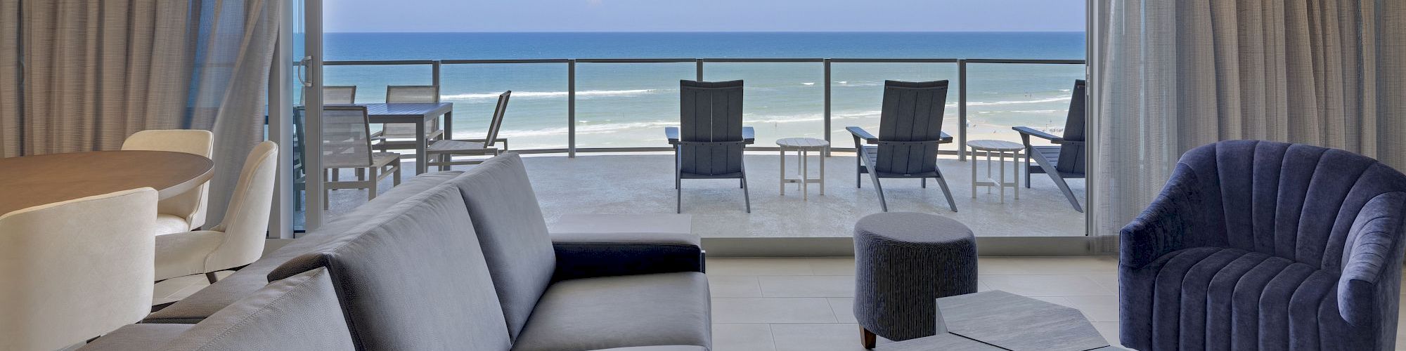 A modern living room with a grey sofa, blue armchair, unique coffee table, and balcony overlooking the ocean.