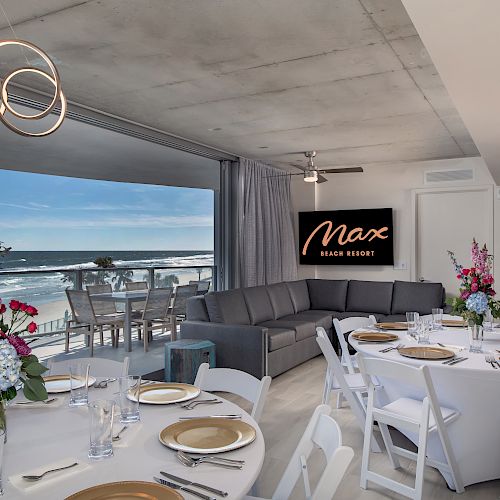 A dining area with round tables set for an event, overlooking a beach view. A couch and sign reading "Max Beach Resort" are in the background.